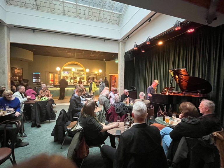 En man spelar på en flygel i ett fint rum med publik vid små bord. I bakgrunden en bardisk. 